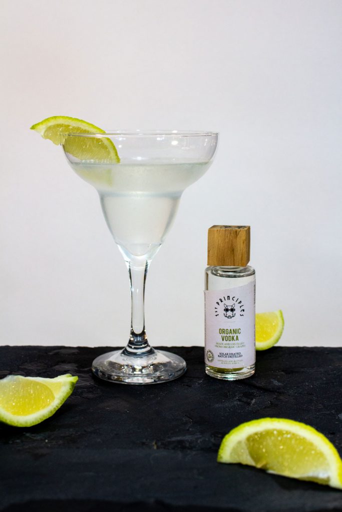 A Gimlet Cocktail made from 1st Principles organic vodka and presented in a margarita cocktail glass with a lemon wedge as garnish.
